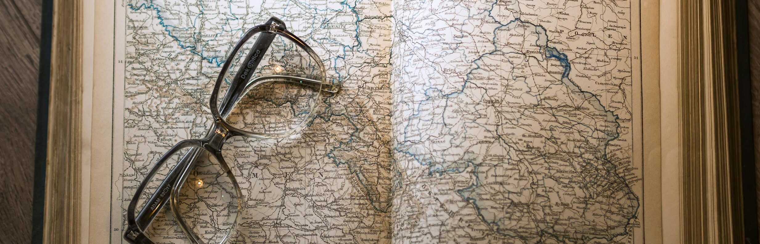 A close-up of a vintage map with eyeglasses resting on an open book, set on a wooden table.